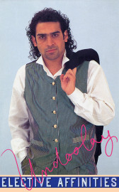 A man in a white shirt and pinstriped vest and pants casually holds a black jacket over one shoulder. 