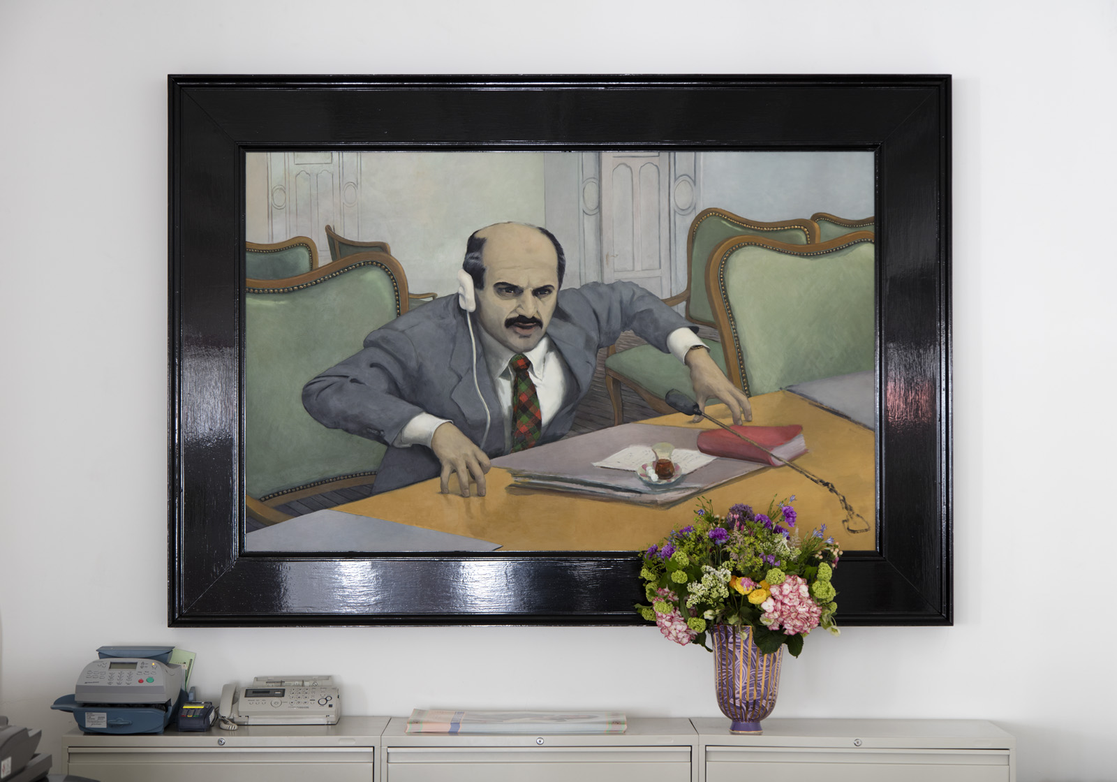 A glossy black frame surrounds a painting of a man in a grey suit seated at a table. The man is wearing a white earpiece and is leaning forward towards a microphone. His fingers are tented on top of the table in an emphatic gesture.