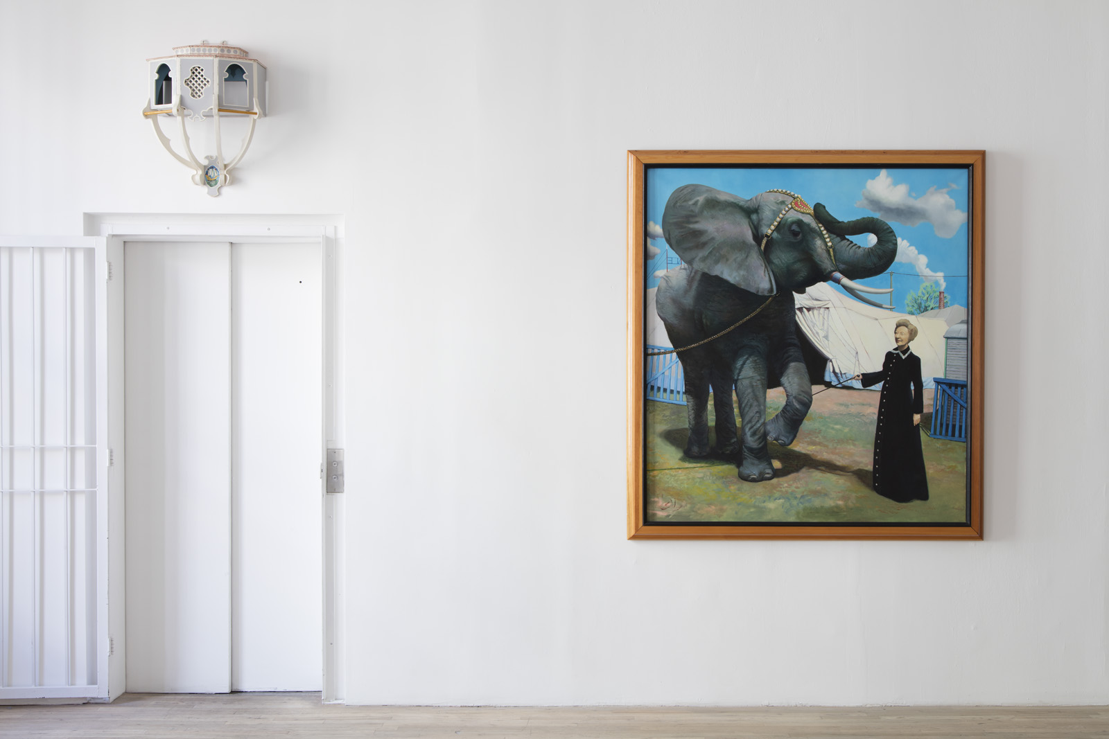 A framed painting is hung on a white wall adjacent to an elevator with a decorative sconce installed above the painted white doors. The painting depicts a chained elephant being trained by a woman in a black dress. The woman holds a baton against the elephant's raised foreleg.
