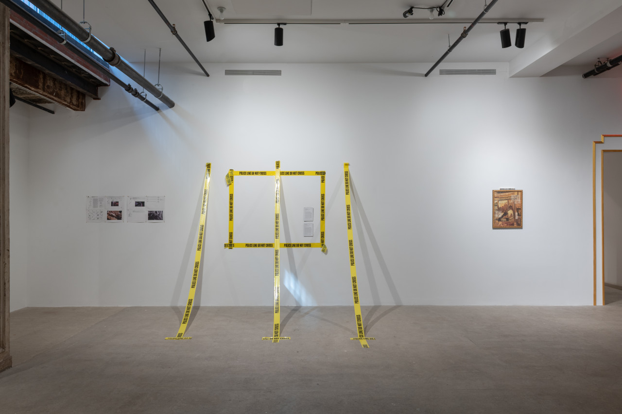 A color photograph of a gallery space. Hanging from the wall is a printer manual, police tape outlining a rectangle with three additional strips extending from the wall to the floor, and a wooden rectangular painting.