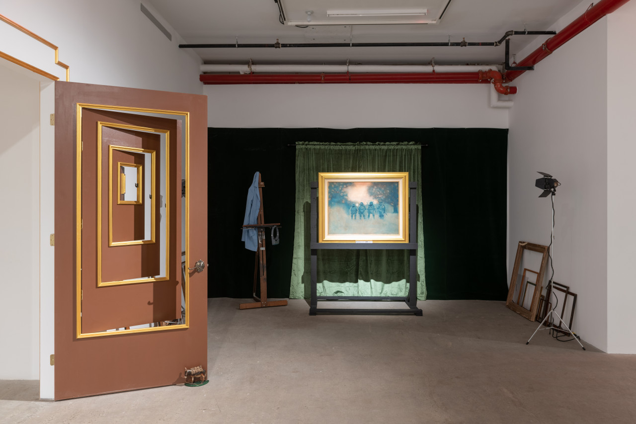 A color photograph of a door with multiple nested smaller doors within the doorframe. Each individual doorway is framed with a gold outline. To the left are several empty picture frames and two studio lights. In front of dark green curtains is a painting set in a gold frame.