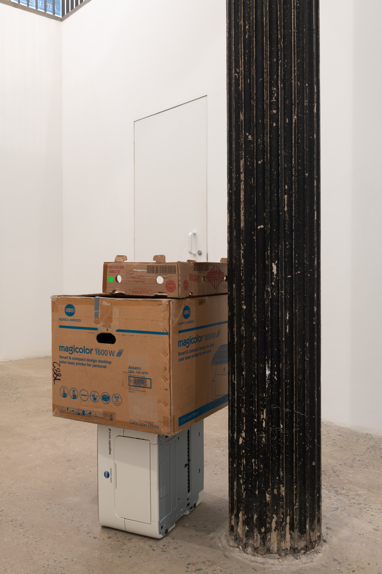 A color photograph of two cardboard boxes stacked on a printer and resting against a dark column in a gallery space. The top box is narrow and open at the top, and the second box is large with blue text and detailing.