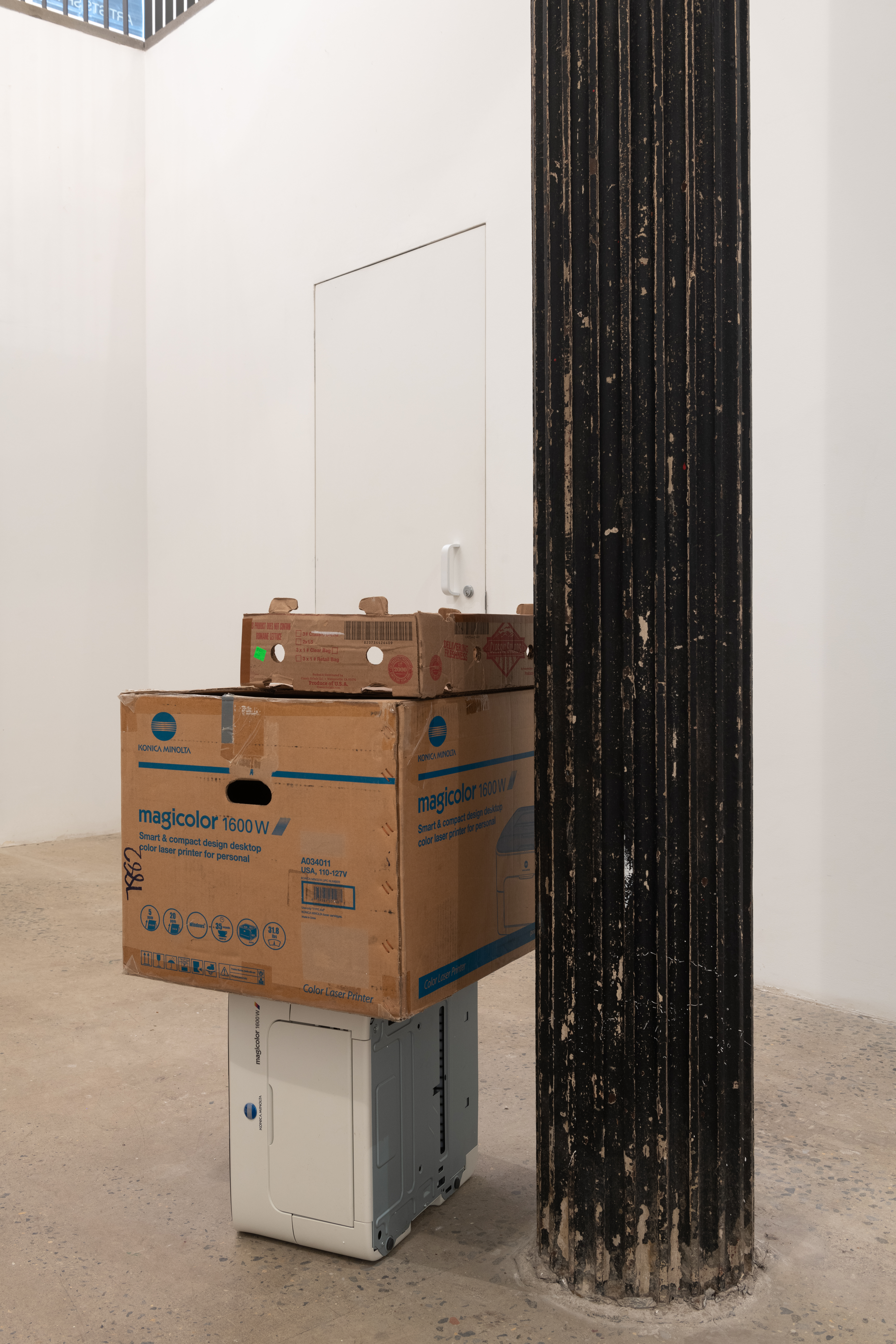 A color photograph of two cardboard boxes stacked on a printer and resting against a dark column in a gallery space. The top box is narrow and open at the top, and the second box is large with blue text and detailing.