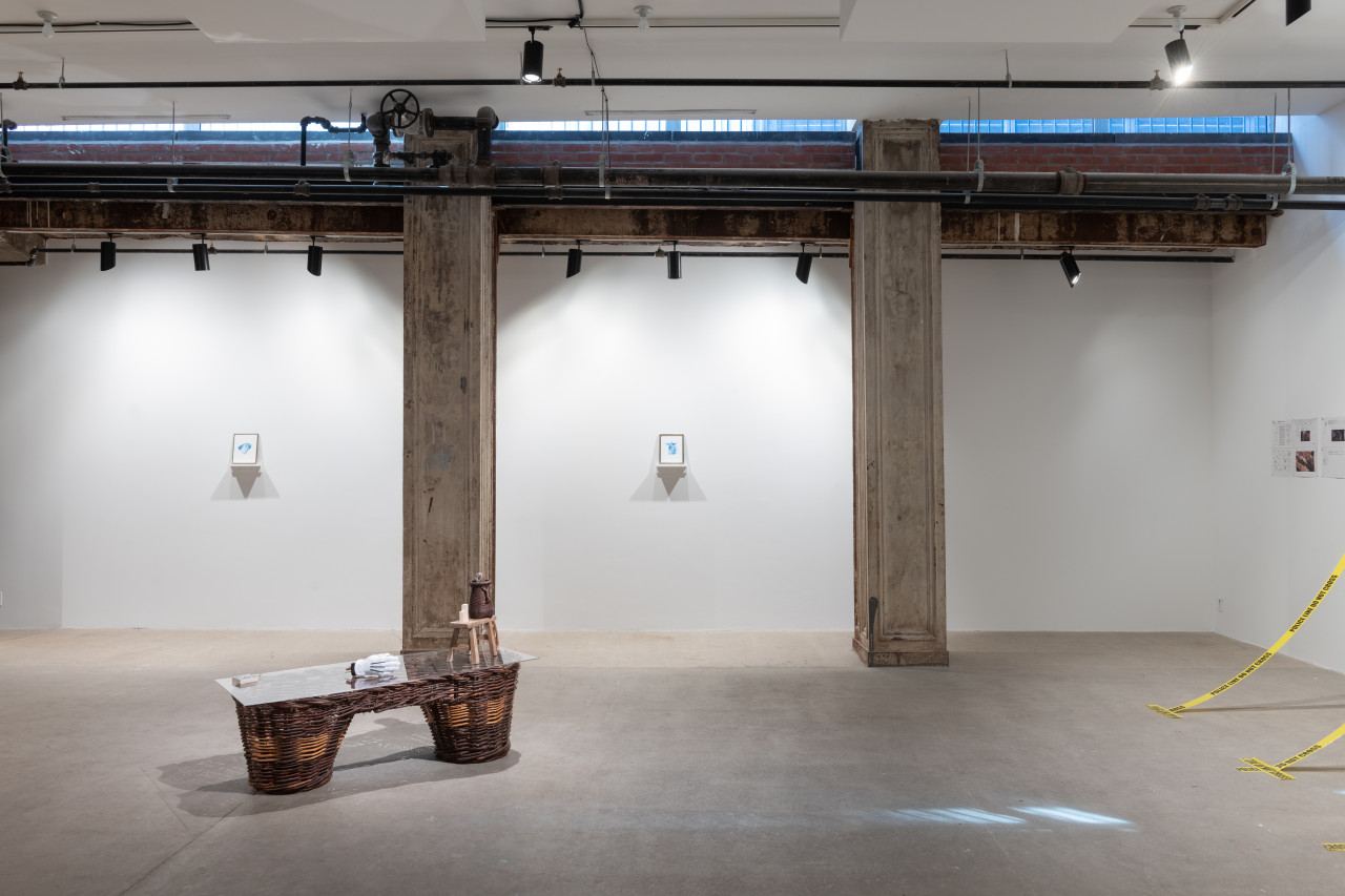 A color photograph of a gallery space. Two small paintings hang on the wall between concrete columns set into the wall. In the foreground, a large wicker pannier is visible. Two strips of police tape can be seen in the right corner of the frame.