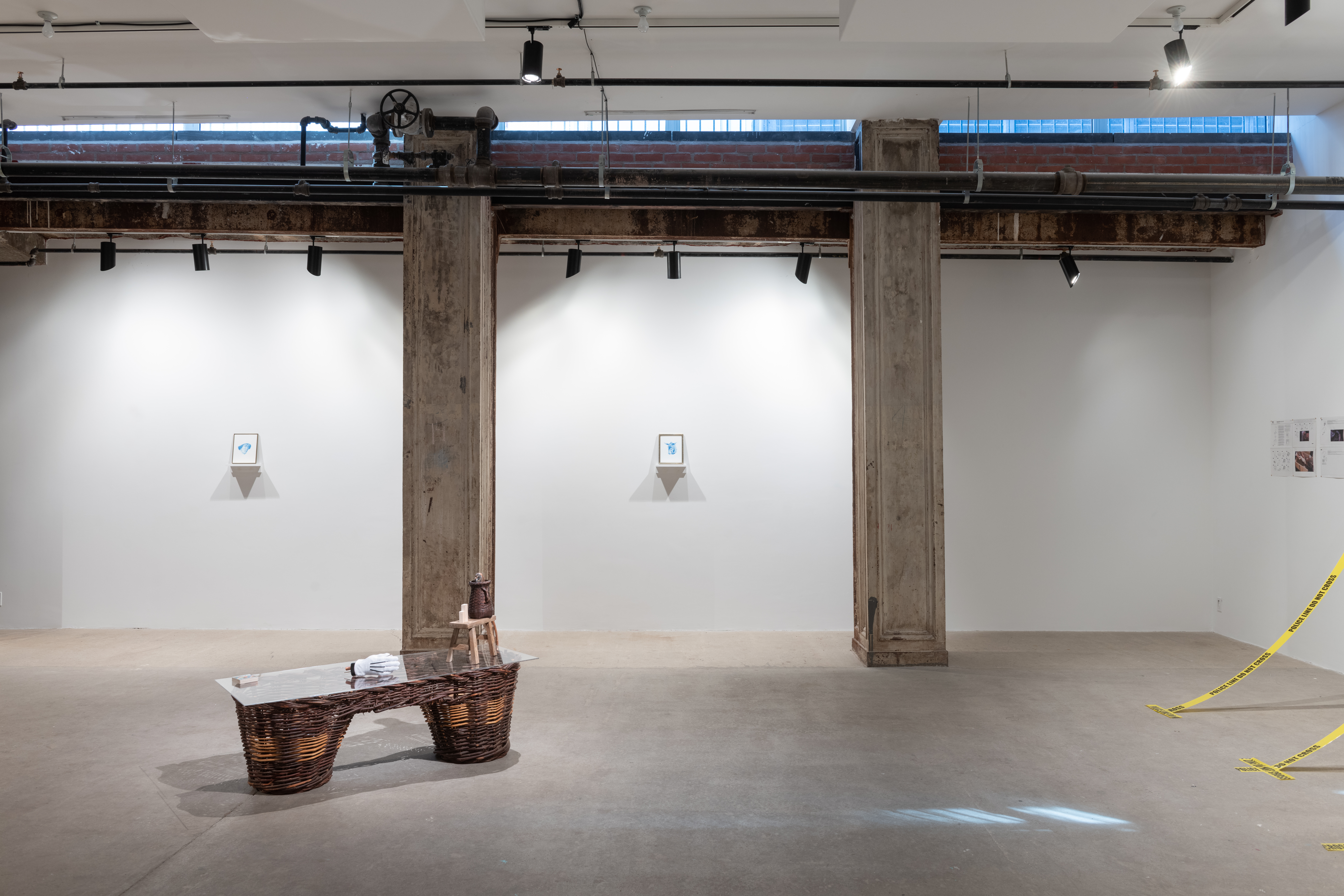 A color photograph of a gallery space. Two small paintings hang on the wall between concrete columns set into the wall. In the foreground, a large wicker pannier is visible. Two strips of police tape can be seen in the right corner of the frame.