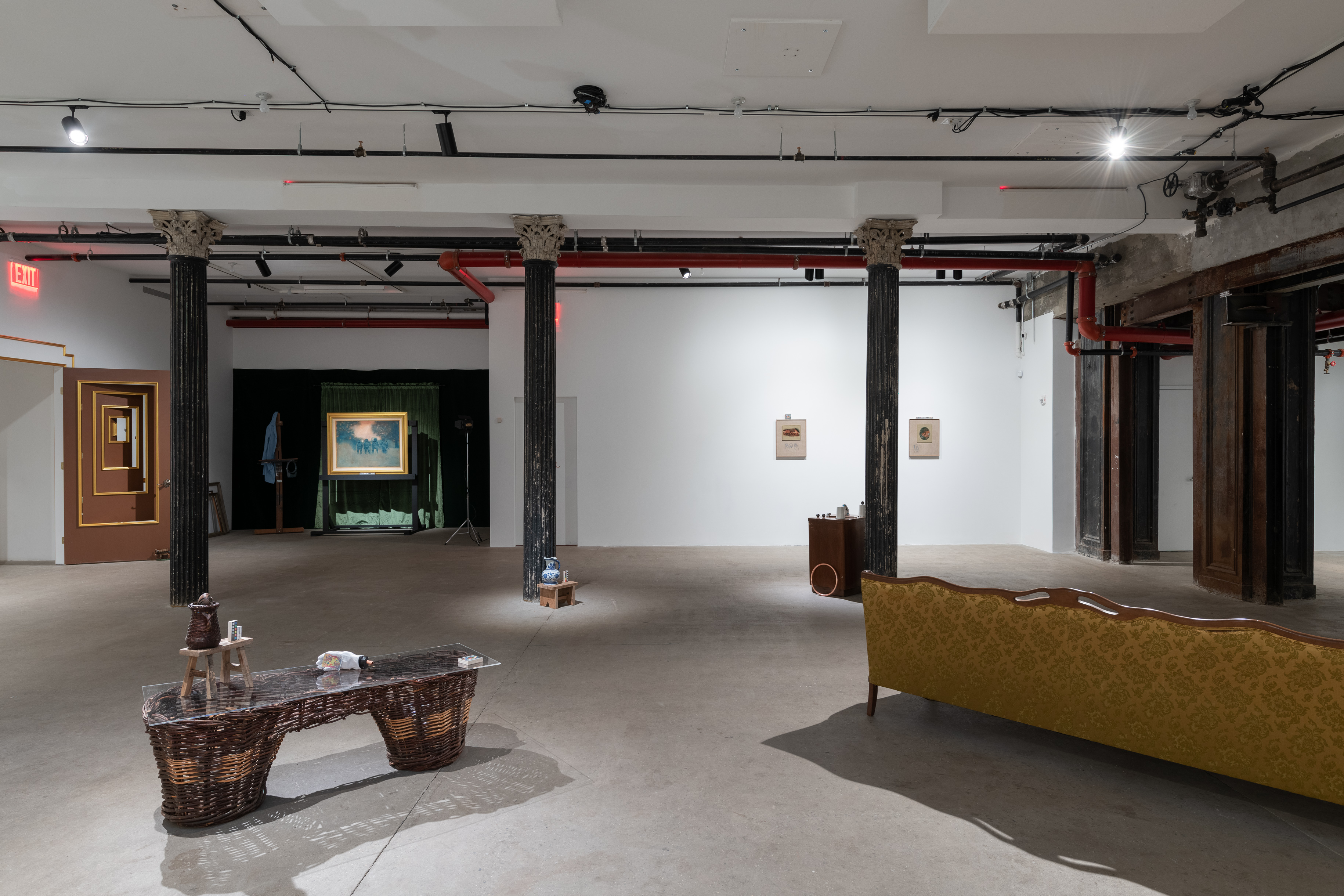 A color installation view of a gallery space with three dark columns. A wicker pannier and a yellow brocade couch are visible in the foreground. In the background, a multi-tiered door, several paintings, a wooden stool and nightstand are also visible.