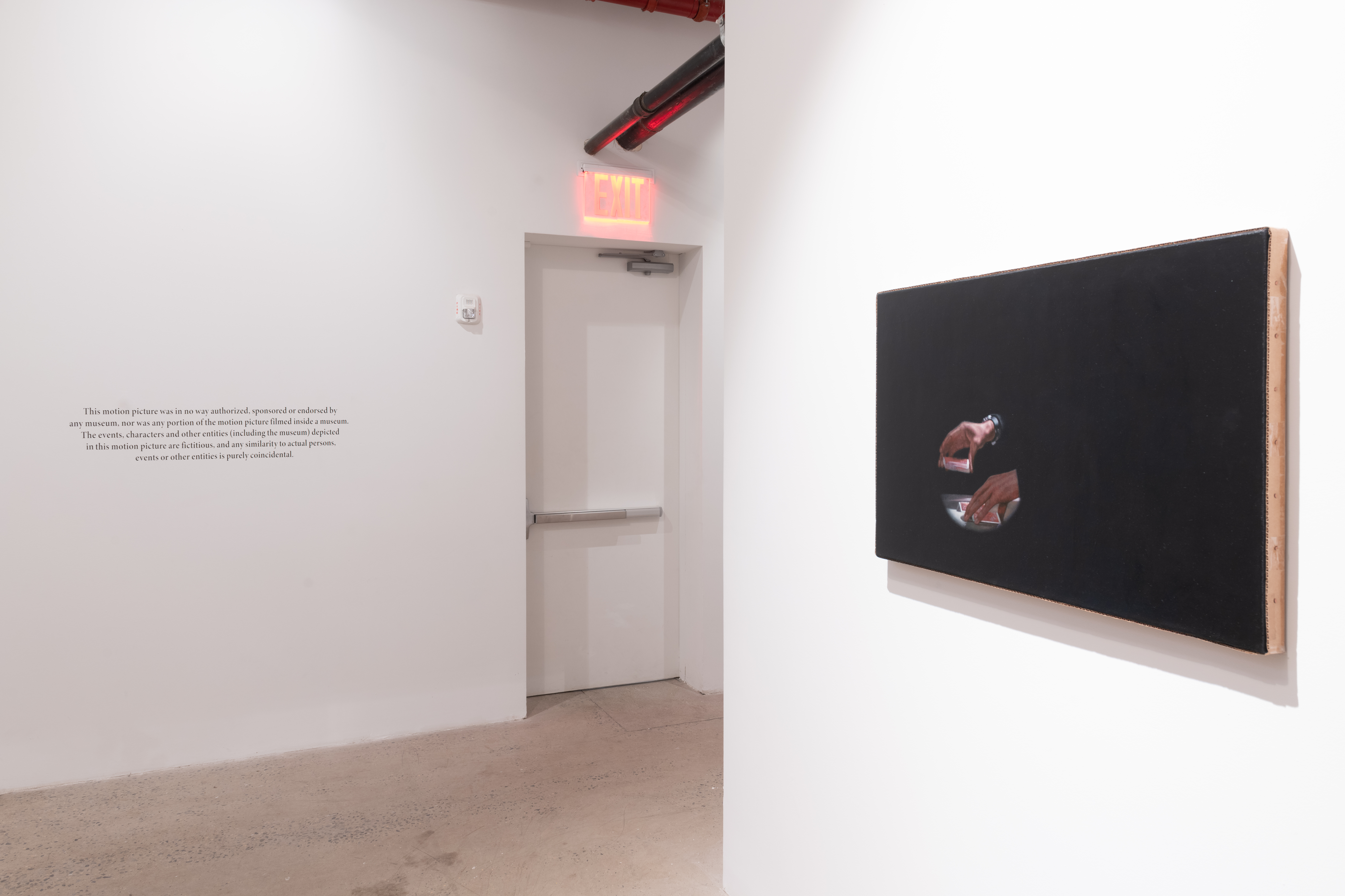 A color photograph of a painting in a gallery space. The canvas is mostly dark, featuring a set of hands with cards in the lower left corner. In the background, some wall text is visible next to a door with a red exit sign above.