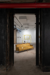 A color photograph of a yellow couch in a gallery space. A cane with a white glove rests on the couch cushions and a rope extends from its base. In the background, police tape demarcates a rectangle on a wall, and three strips of police tape extend from the wall to the floor.