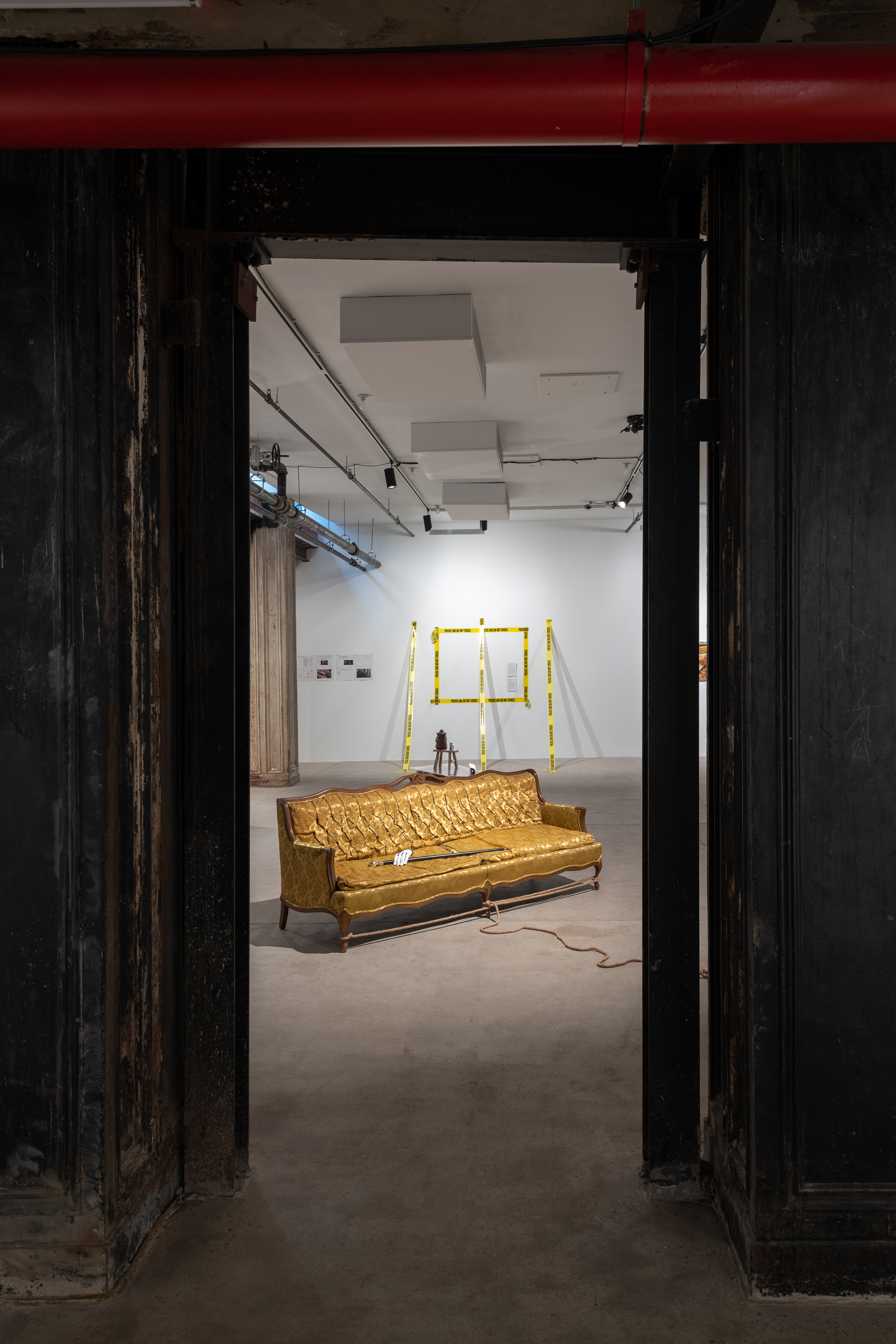 A color photograph of a yellow couch in a gallery space. A cane with a white glove rests on the couch cushions and a rope extends from its base. In the background, police tape demarcates a rectangle on a wall, and three strips of police tape extend from the wall to the floor.