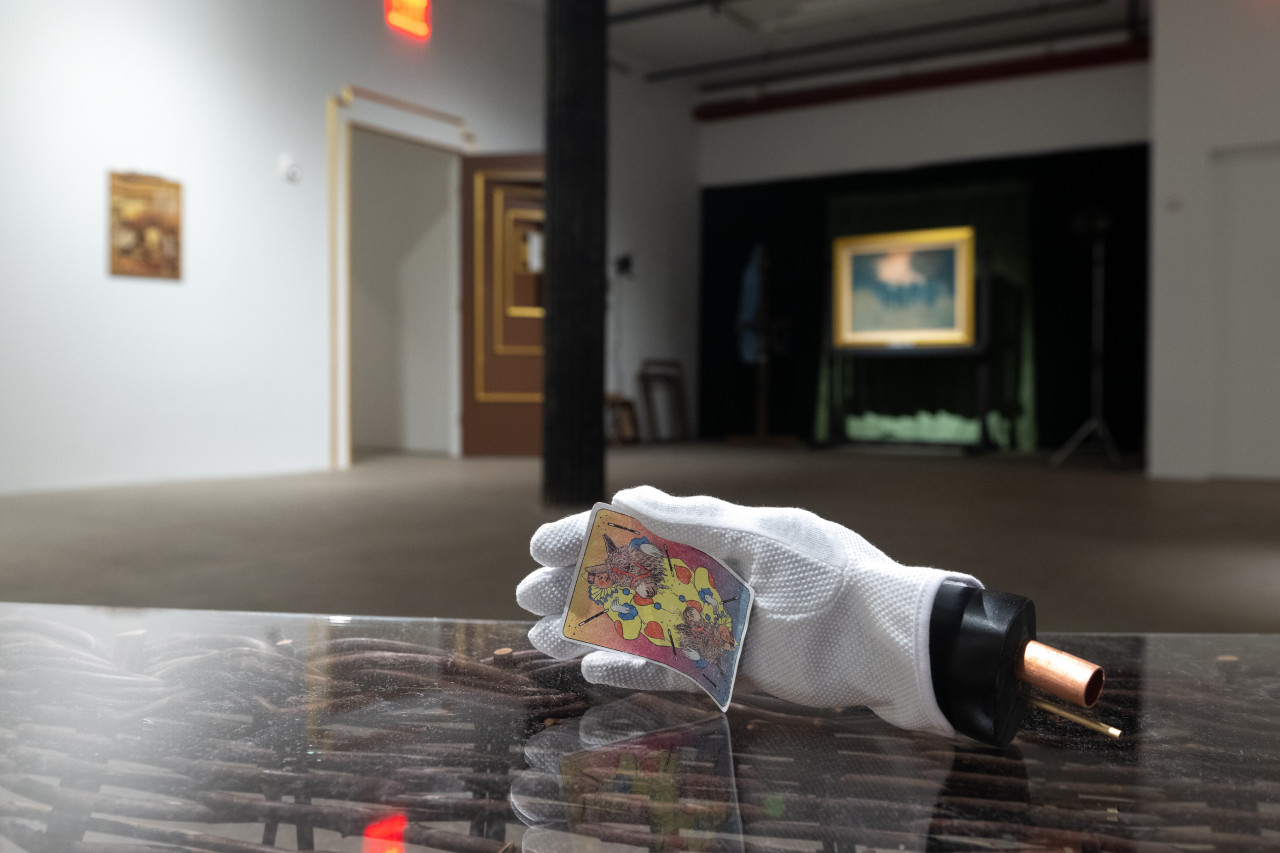 A color photograph of a white glove resting on a wicker pannier in a gallery space. The glove cups a multicolored card. Further in the background, an illuminated painting in a gold frame and a multi-tiered door are visible.