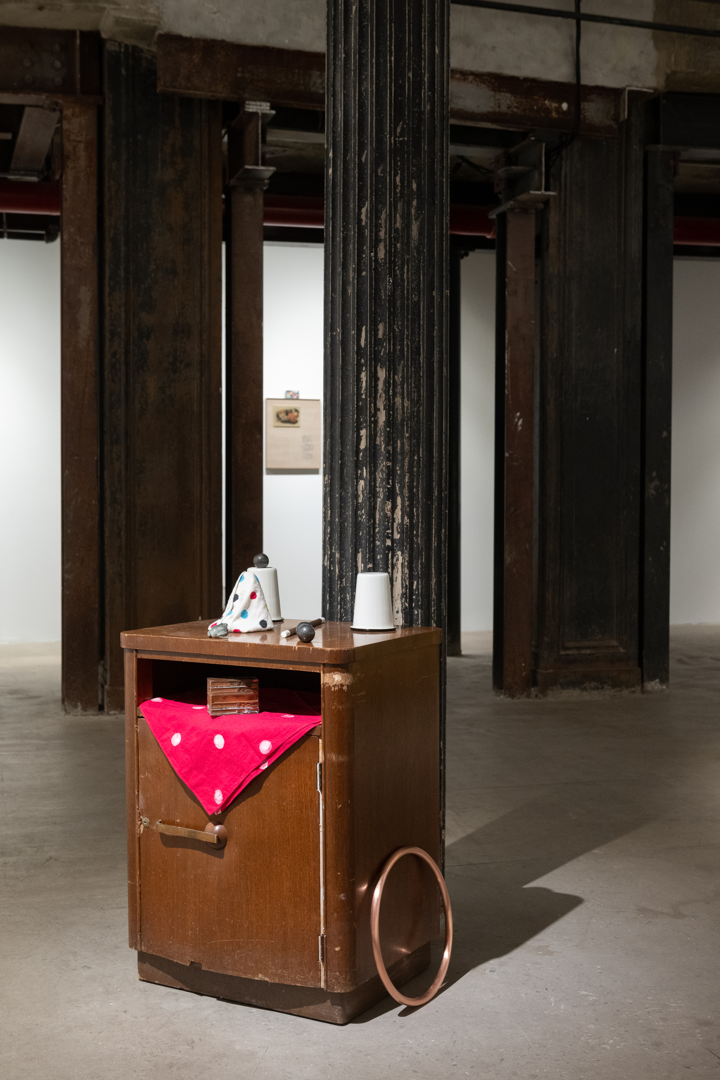 A color photograph of a wooden nightstand with a red hankerchief, ceramic frog, wand, and enamel tumblers. On the right side of the nightstand, a brass hoop rests on the floor, propped up against the nightstand.