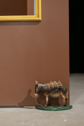 A color photograph of a doorstop shaped like a donkey. The donkey carries a bundle of sticks on its back and props open a brown door.