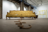 A color photograph of a yellow brocade couch in a gallery space. A long tangled rope extends from the bottom of the couch towards the camera. In the background, police tape against frames a rectangle on the wall, and three additional strips of police tape extend from the wall to the floor.