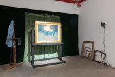 A color photograph of an installation. In front of dark green curtains, a painting in a gold frame is illuminated. To the left, a conservator's coat hangs from an easel. Several empty frames and a studio light are visible on the right.