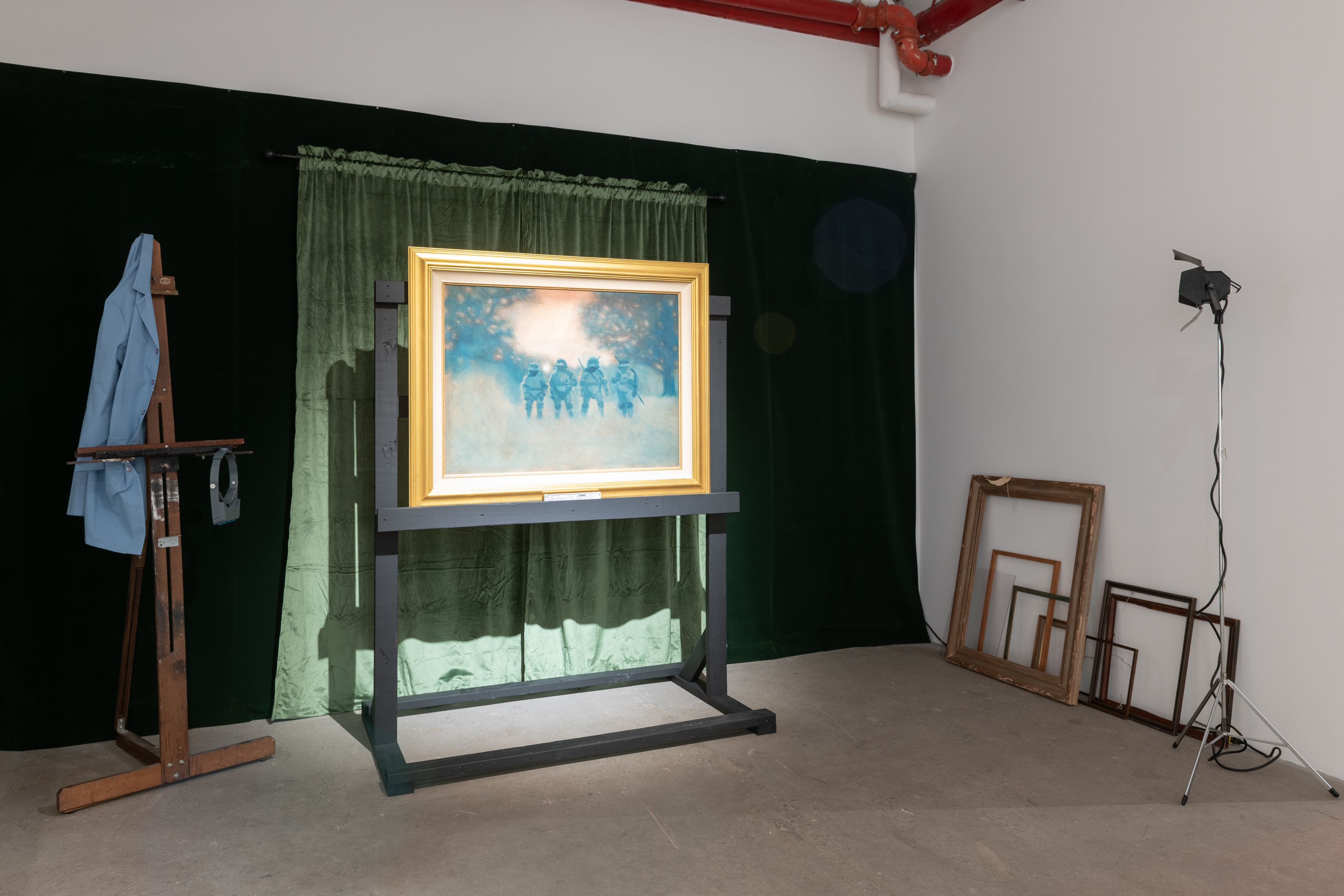 A color photograph of an installation. In front of dark green curtains, a painting in a gold frame is illuminated. To the left, a conservator's coat hangs from an easel. Several empty frames and a studio light are visible on the right.