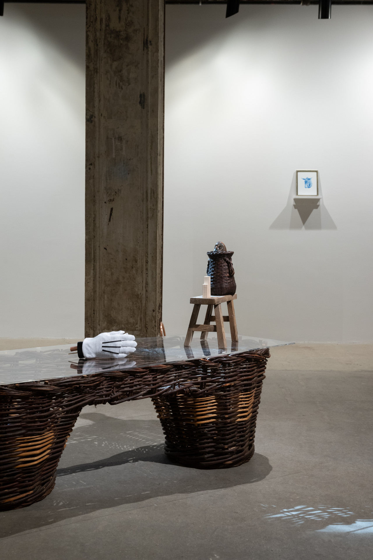 A color photograph of a gallery space. In the foreground is a large wicker pannier with a white glove, small wooden stool, and ceramic jug on top. In the background a small portrait of a donkey in blue is visible.