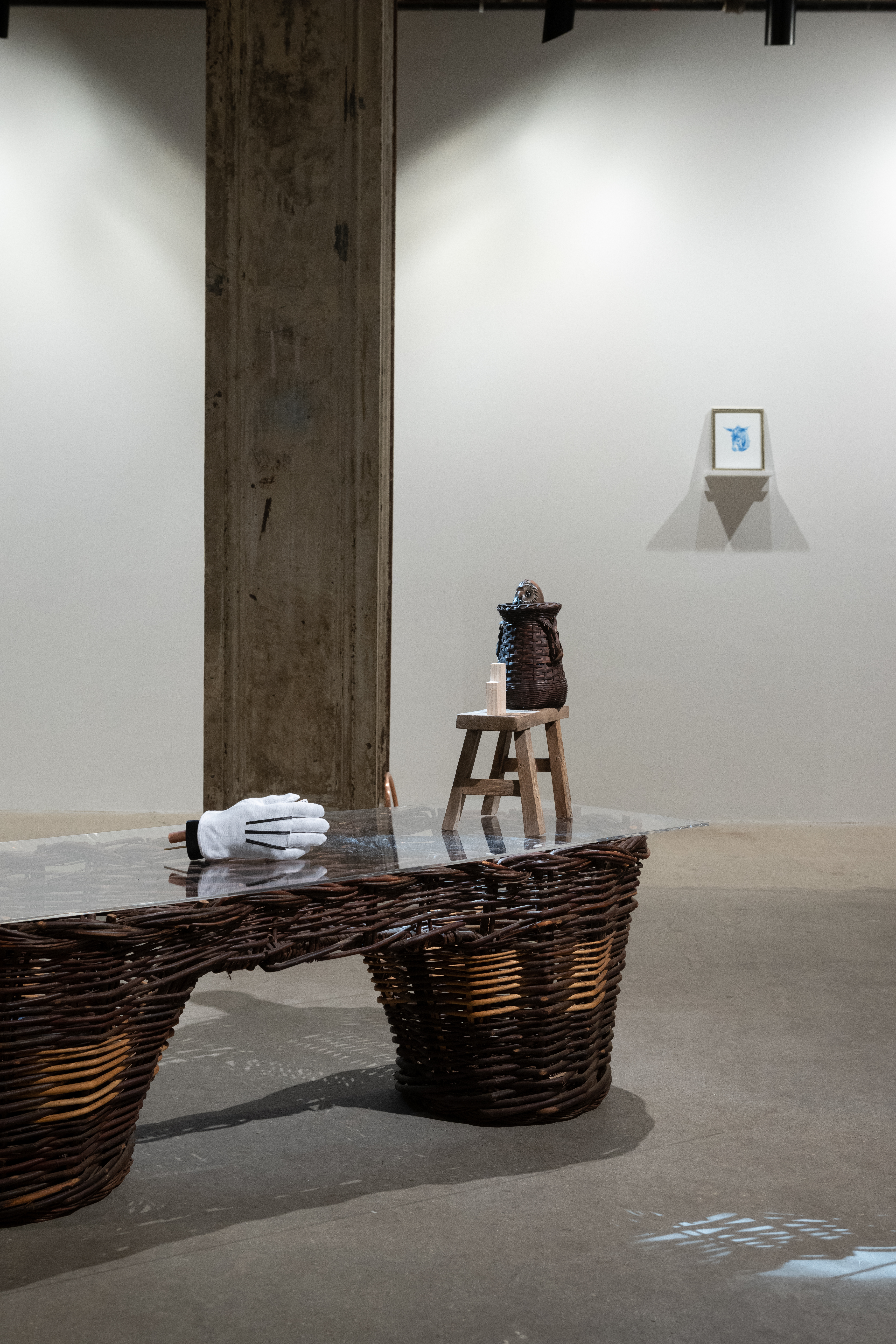 A color photograph of a gallery space. In the foreground is a large wicker pannier with a white glove, small wooden stool, and ceramic jug on top. In the background a small portrait of a donkey in blue is visible.