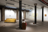 A color installation view of a gallery space with several dark columns. A yellow brocade couch, wooden nightstand, wicker pannier, and police tape outlining a rectangle on the wall with three additional strips extending from the wall to the floor are visible.