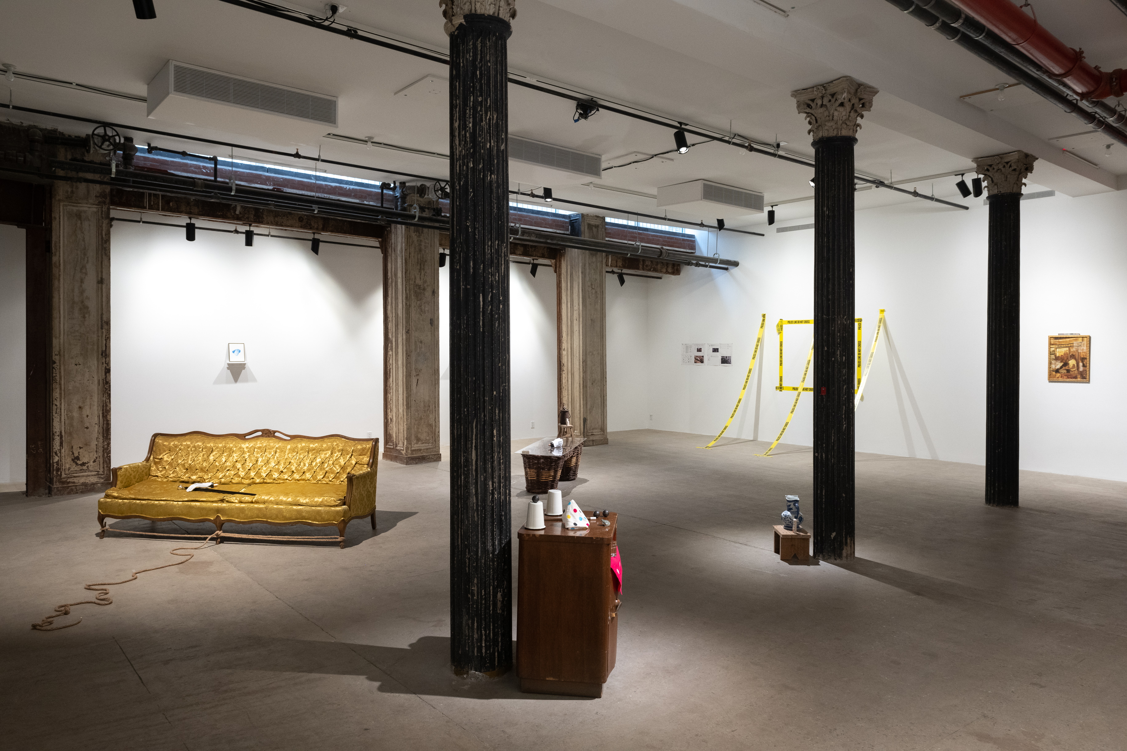 A color installation view of a gallery space with several dark columns. A yellow brocade couch, wooden nightstand, wicker pannier, and police tape outlining a rectangle on the wall with three additional strips extending from the wall to the floor are visible.