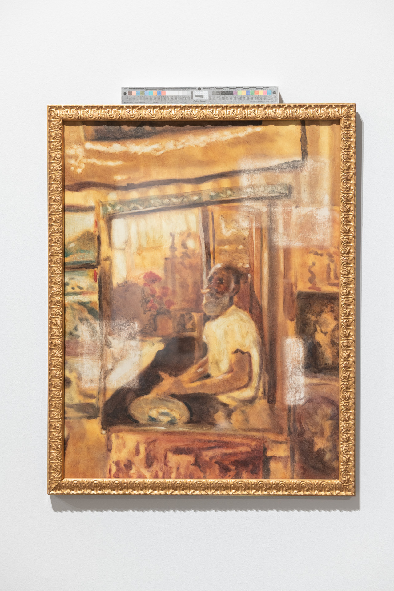 A gold framed painting of a seated figure with a grey beard in a tawny toned space.