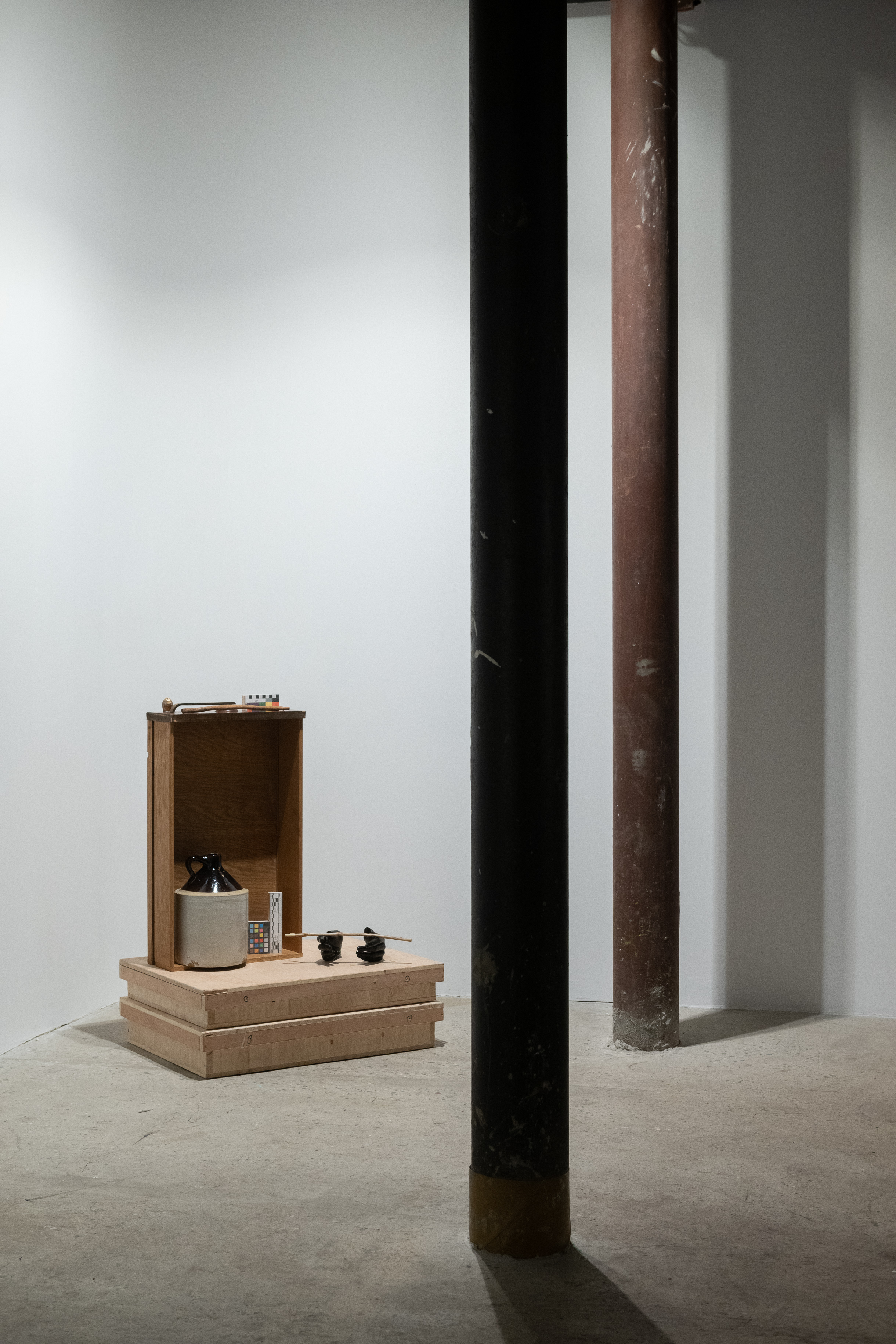 A color photograph of a drawer positioned vertically on plywood in the corner of a gallery space. In front of the drawer is a ceramic jug and several other obkects including a wand.