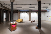 A color photograph of a gallery space with several dark columns. A small wooden chest with a red piece of fabric and three white objects on top along with a small wooden stool are visible in the foreground. Further in the background are a yellow couch and a large wicker pannier..
