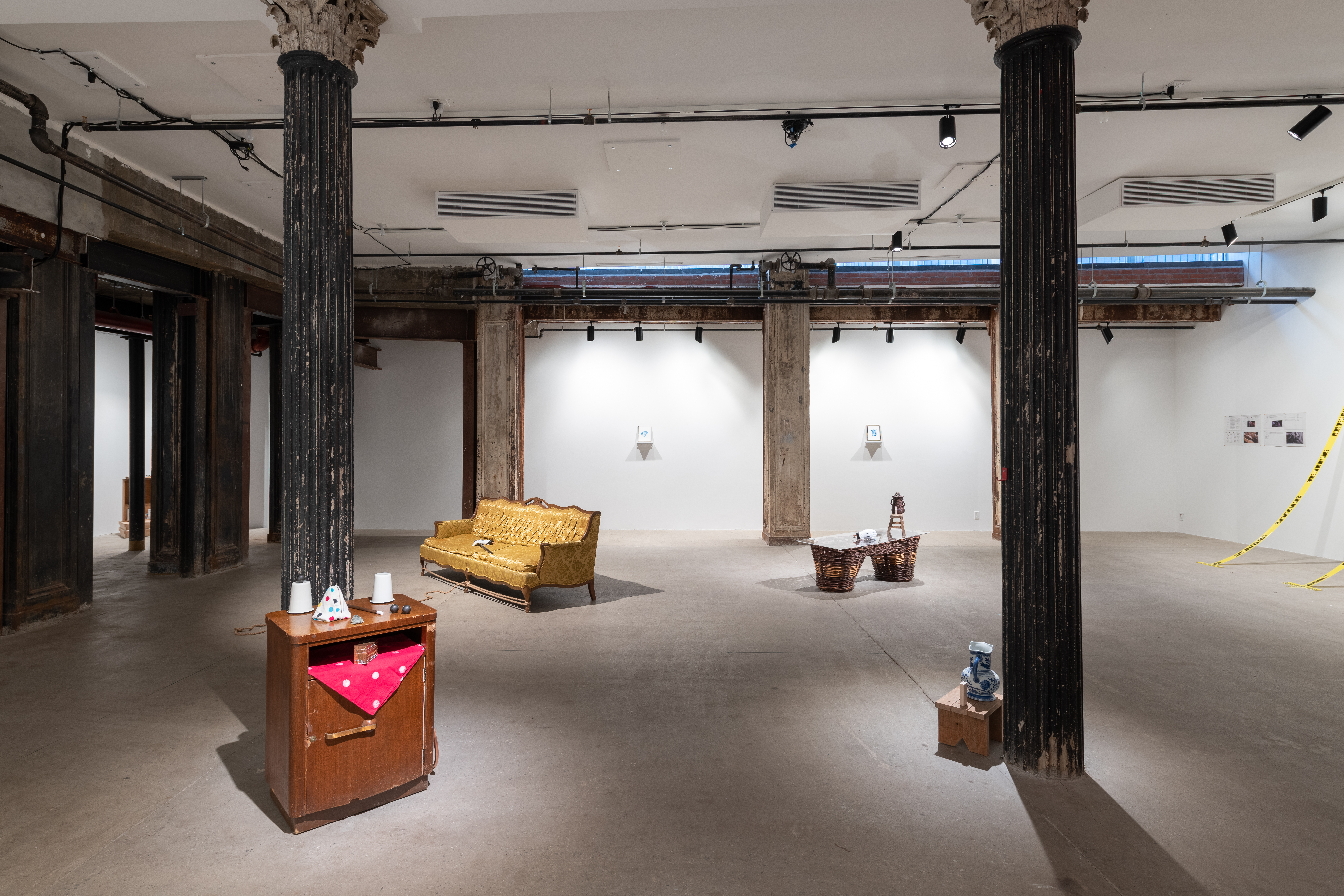 A color photograph of a gallery space with several dark columns. A small wooden chest with a red piece of fabric and three white objects on top along with a small wooden stool are visible in the foreground. Further in the background are a yellow couch and a large wicker pannier..