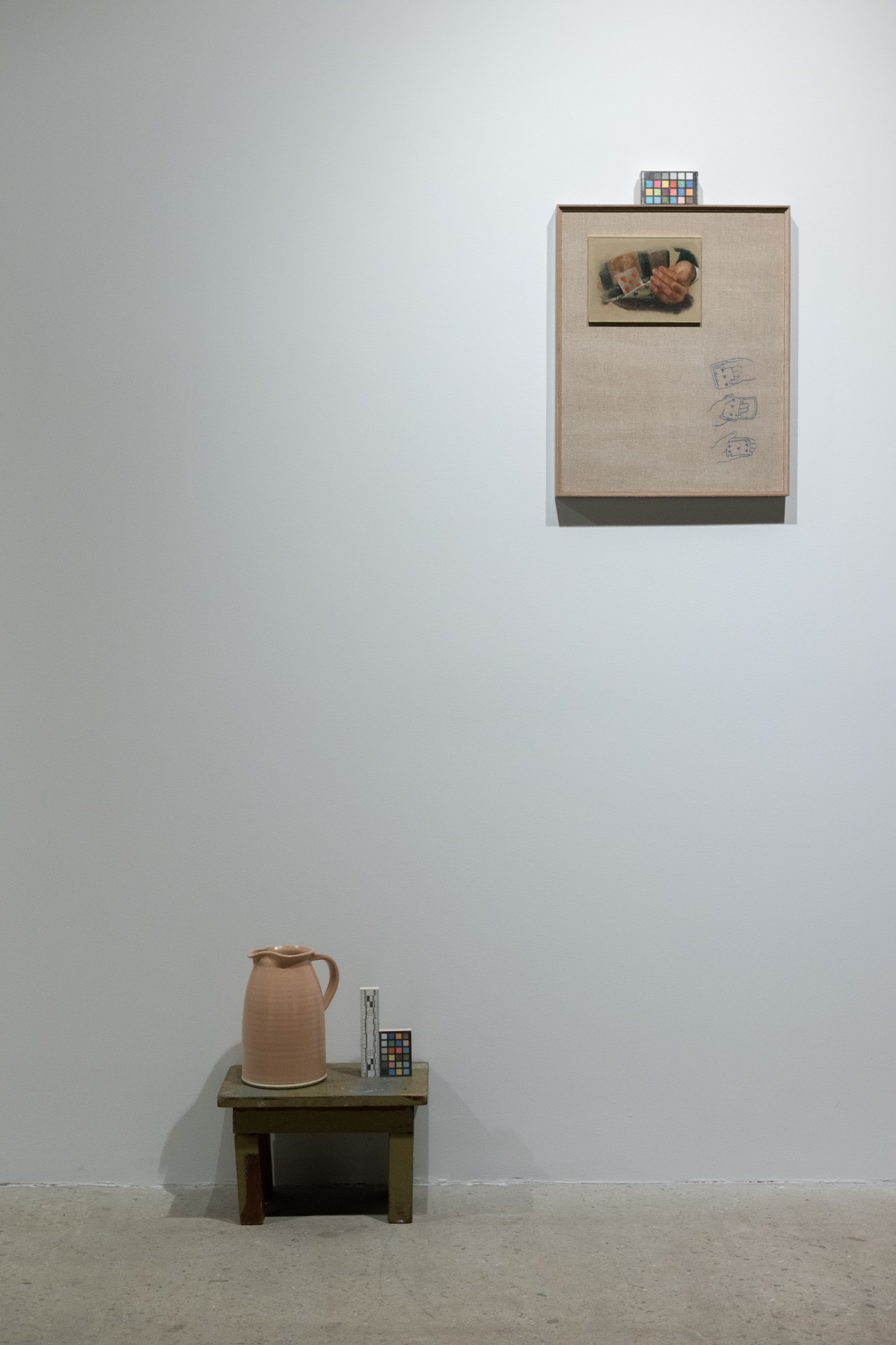 A color photograph of a ceramic jub resting on a small wooden stool next to a color control strip. On the wall above and to the right is a painting of two hands. Another color control strip rests on top of the painting