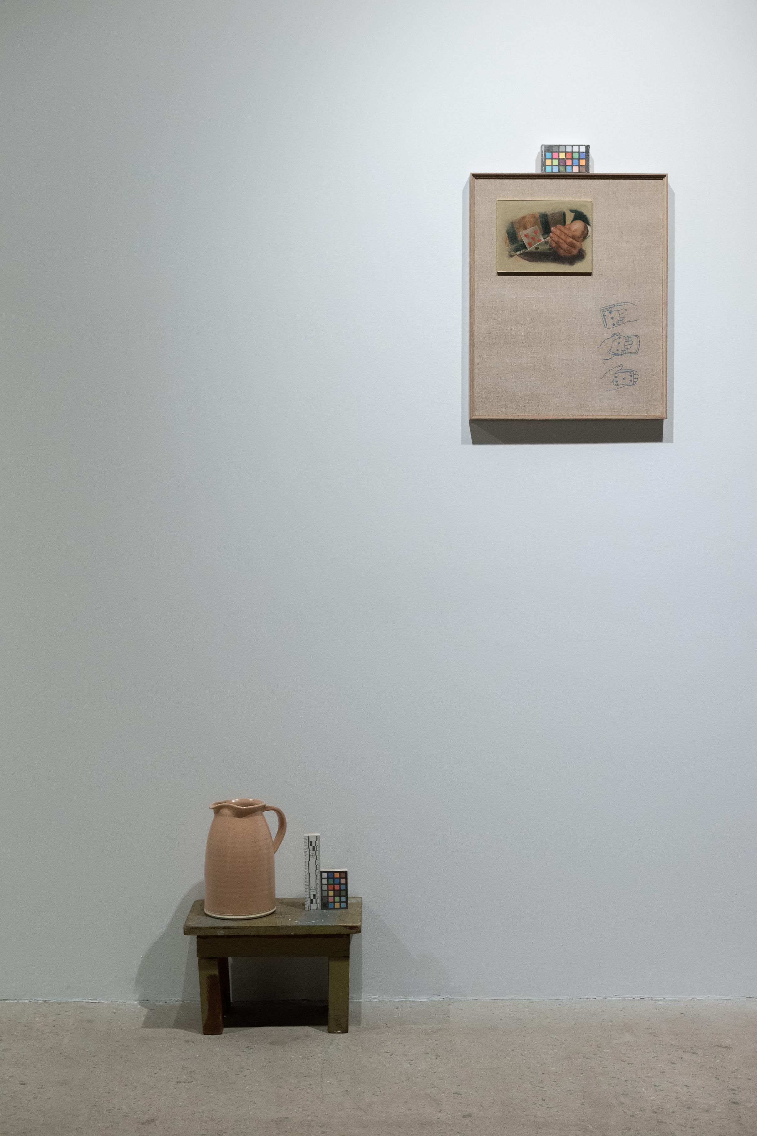 A color photograph of a ceramic jub resting on a small wooden stool next to a color control strip. On the wall above and to the right is a painting of two hands. Another color control strip rests on top of the painting's frame.