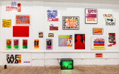 Many colorful documents, photographs, and posters installed on a white wall in an exhibition space. A small television monitor displaying a green image sits on the ground beneath the posters and other artworks.