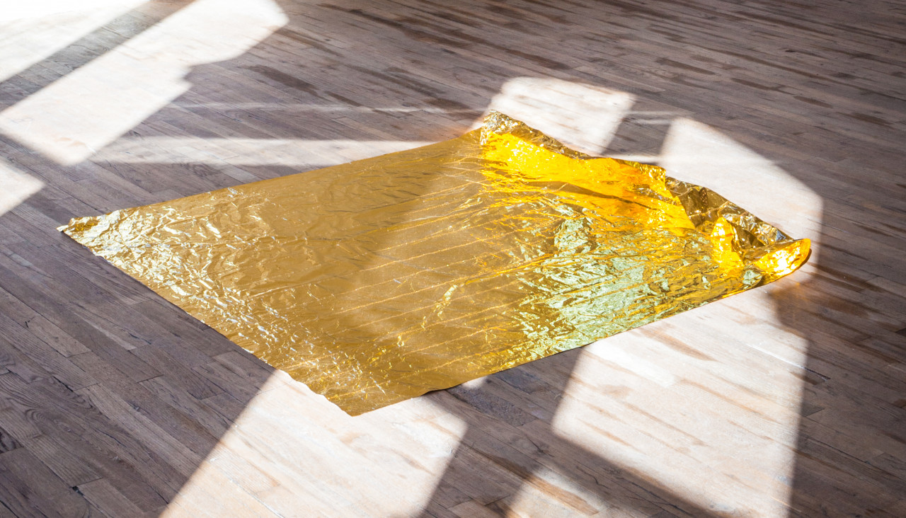 A rectangle of gold foil laying flat on a wooden floor. Light descending from a nearby window illuminates the foil.