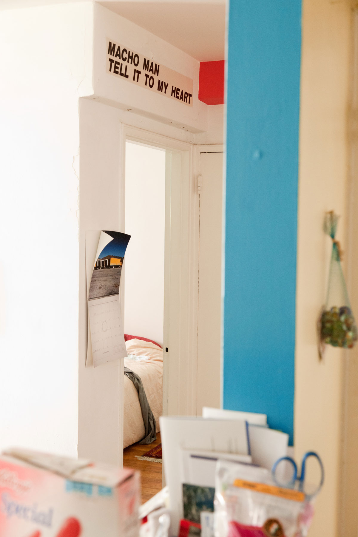 An angled view from a ledge containing household items, into a bedroom. Above the doorway black text reads, 