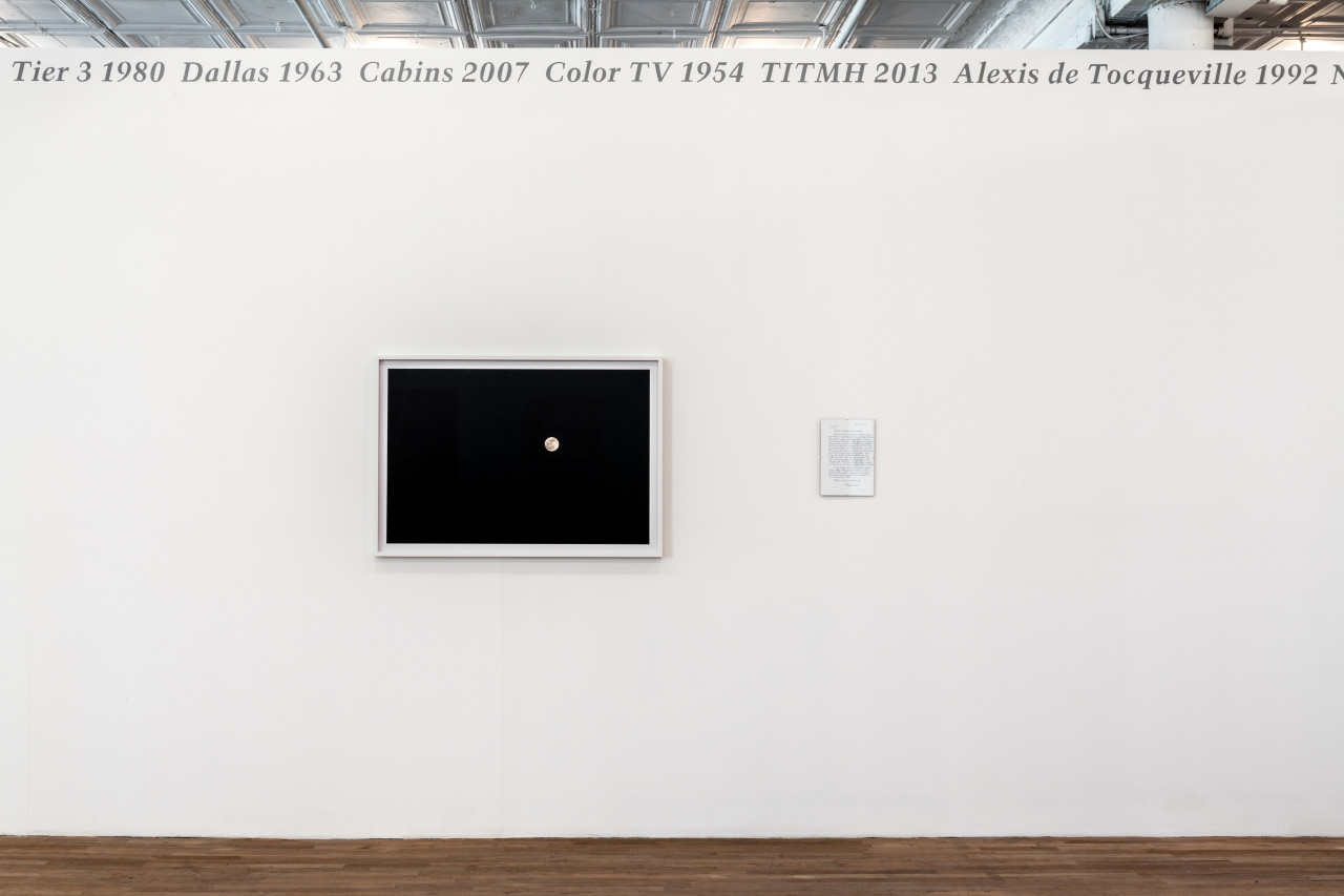 Silver, italic text displayed across the top edge of a white gallery wall reads, "Tier 3 1980 Dallas 1963 Cabins 2007 Color TV 1954 TITMH 2013 Alexis de Tocqueville 1992." An image of the moon in a black sky and a small letter are displayed side-by-side on the gallery wall beneath the silver text.