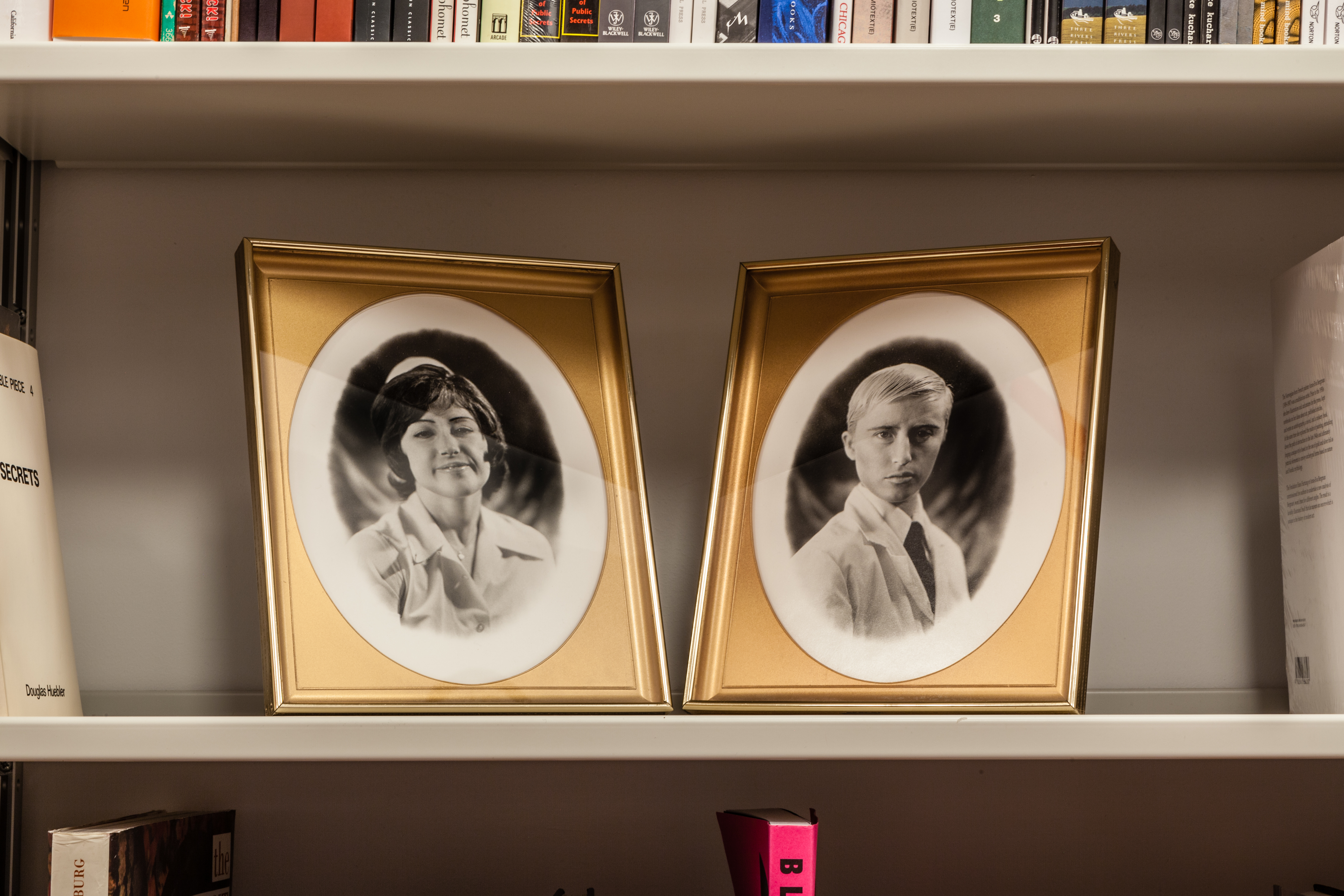 Two gold picture frames with black and white portraits of a woman (left) and a man (right) face each other on a shelf.