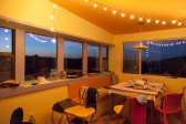 A warm colored room, surrounded by windows across two walls with view to a dimly lit horizon. String lights illuminate a scene of miscellaneous items scattered across a living space furnished with a table and chairs.