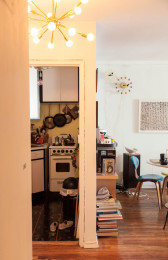 A portrait-oriented view of a home interior. On the left, a doorway leads to a kitchen. On the right, the room opens to a living space with stacks of books on the floor and decor on the wall.