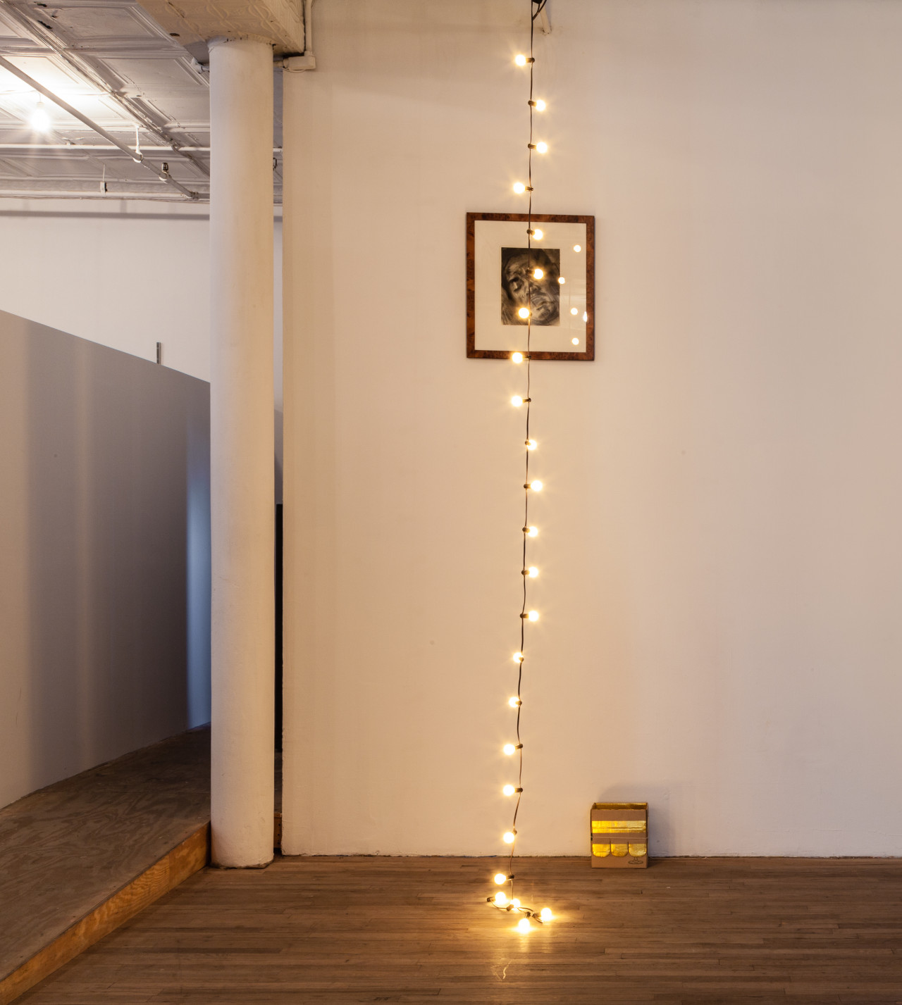A string of light bulbs decends from the celing of an exhibition space, with the end of its wiring resting on the ground below. A framed image of a figure