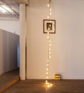 A string of light bulbs decends from the celing of an exhibition space, with the end of its wiring resting on the ground below. A framed image of a figure's face is installed high on the wall directly behind the light string. A small cardboard box with gold details sits on the ground to the right of the light string.