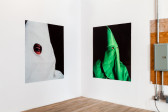 Two white walls forming a corner in an exhibition space. Displayed on the left wall is a large a photograph of an eye visible through a round hole in a white fabric hood. Displayed on the right wall is a photograph of a figure in a green KKK hood against a black background.