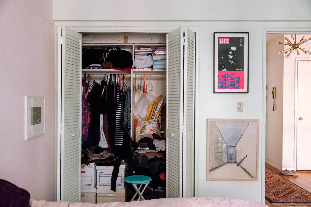 A closet, sliding doors opened to reveal clothing folded on shelves and organized on hangers. To the right, a view into the hallway.