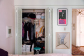 A closet, sliding doors opened to reveal clothing folded on shelves and organized on hangers. To the right, a view into the hallway.