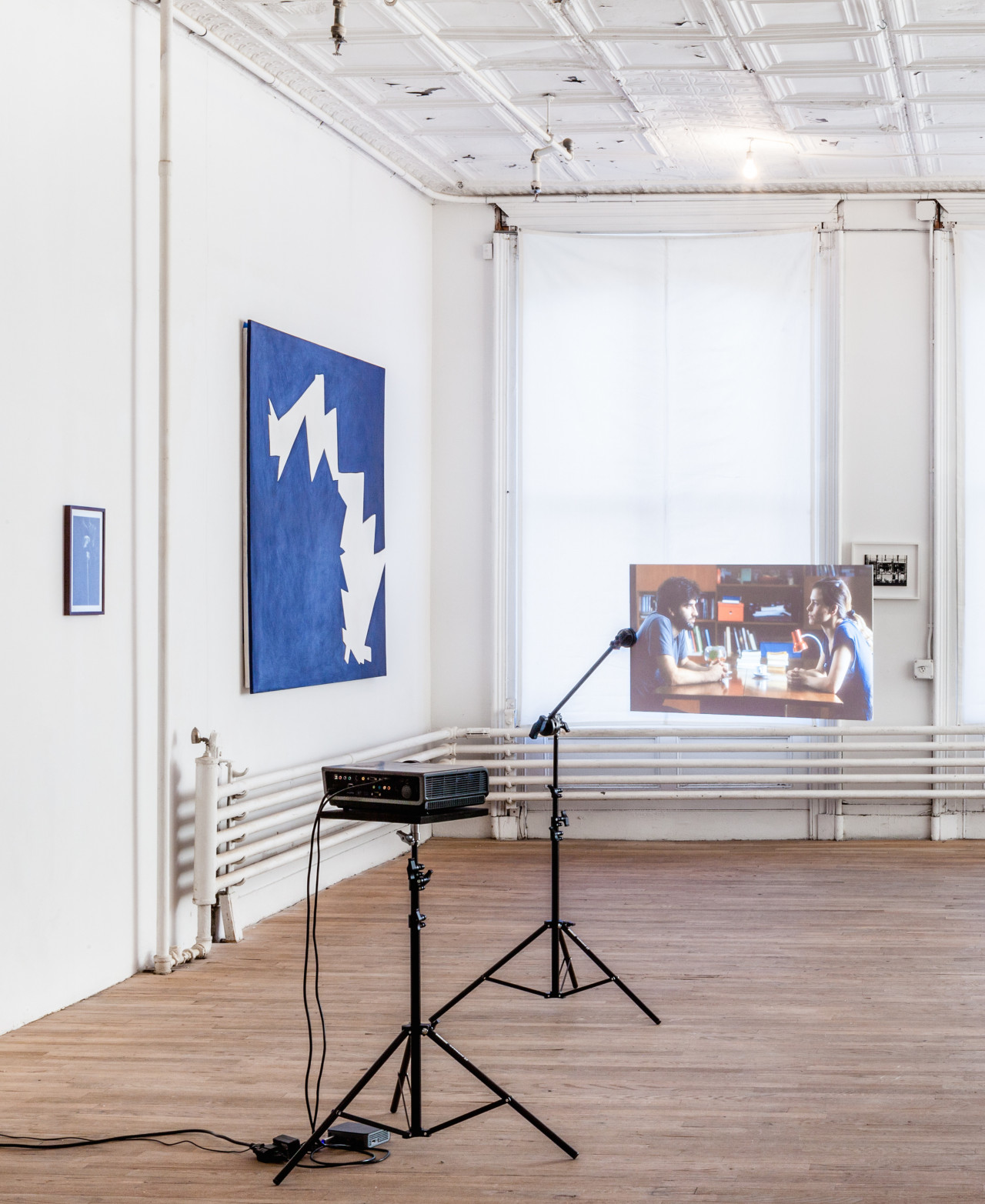 An angled view of large blue painting hanging on a white wall in an exhibition space. To the right of the painting in the center of the room, a projector positioned on a small stand projects an image onto a screen that is also supported by a similar, small stand. The projected image depicts a side view of a man and woman sitting across a table.