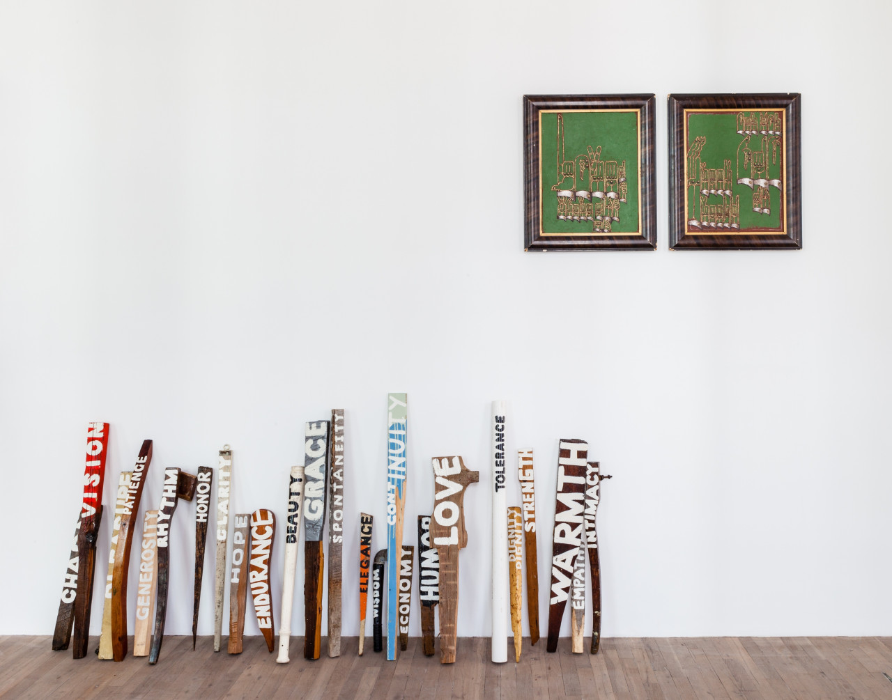 Several small planks of wood of different sizes, shapes, and colors lean against a white gallery wall. Different words have been painted across the length of each plank, including "rhythm," "grace," "love," and "warmth." Above the planks hang two framed, green paintings.