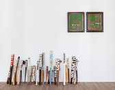 Several small planks of wood of different sizes, shapes, and colors lean against a white gallery wall. Different words have been painted across the length of each plank, including 
