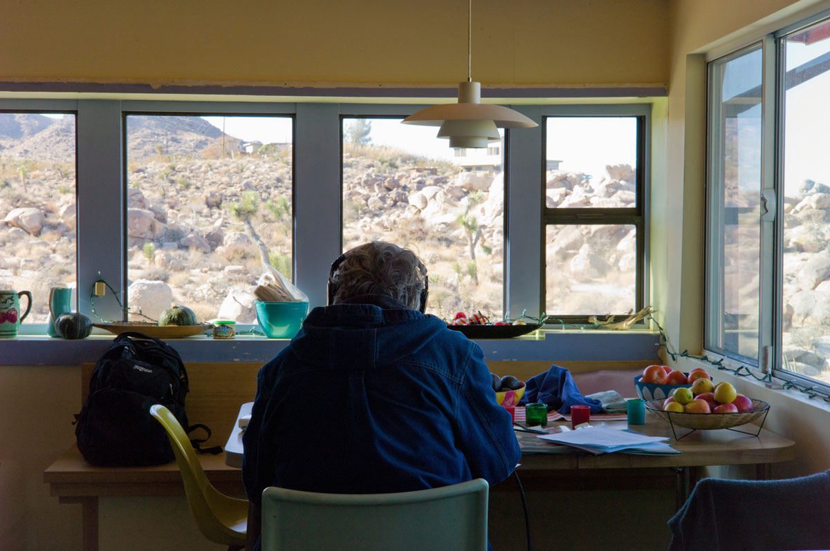 A person wearing a blue jacket and headphones sits at a table, surrounded by windows with view to a rocky, dry terrain.