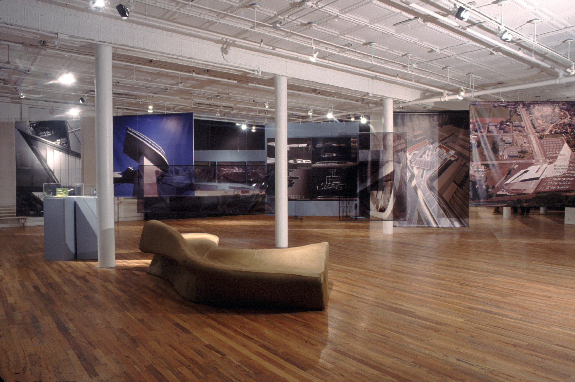 A dimly lit gallery space with wooden flooring, white walls, and white columns. A large, amorphous brown structure with curved sides and a smooth surface is displayed on the gallery floor closest to the viewer. Behind this structure, three columns run diagonally across the gallery space. A gray plinth is located behind the leftmost column and supports several small models. Further back behind the columns and stretching out across the wide gallery space are various photographs of a variety of buildings and building complexes. These images are screenprinted on sheets of tarp that hang from the gallery ceiling.