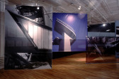 A dimly lit gallery space with wooden flooring. Three large, rectangular photographs of various buildings’ architectural details are suspended from the ceiling, hanging on thin wires. Two of the images have been printed on a tarp-like material, while the image on the far left is suspended within a large sheet of transparent plexiglass.