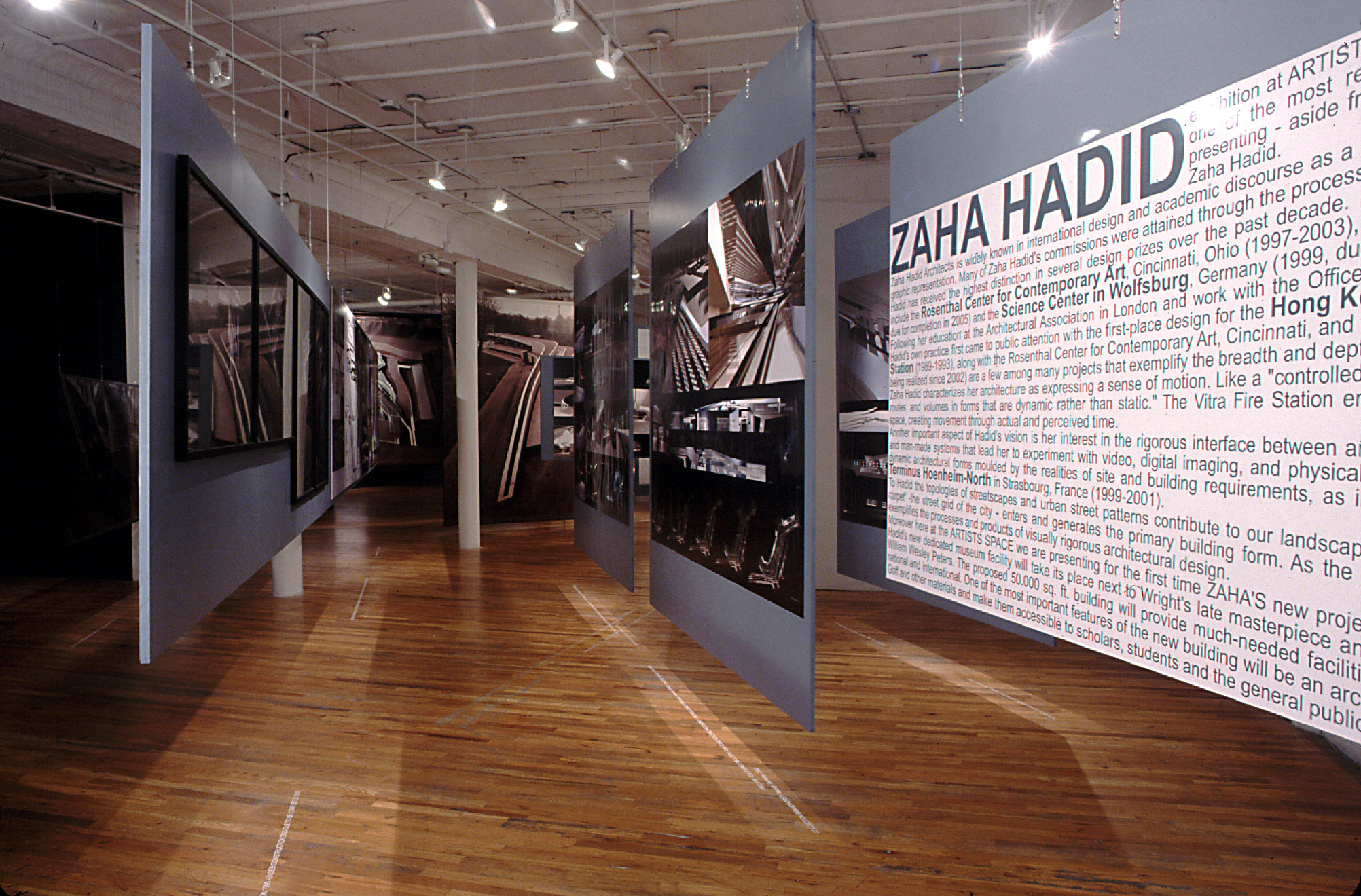 A dimly lit gallery space with wooden flooring. Several sheets of thin gray board hang from the gallery ceiling by thin wires such that they form a set of suspended display walls. The boards are arranged such that they face one another and form a hallway-like area that one can walk through. Various photographs and architectural renderings of buildings are displayed on the walls. A wall arranged to the far right of the others displays black san-serif wall text. Large header text on the wall reads, “ZAHA HADID.”