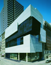 Photograph of a large building on an urban street composed of rectangular sections of dark black and white stone that jut variously in towards and away from the building's facade. The bottom portion of the building is supported by two white columns and has a glass facade.