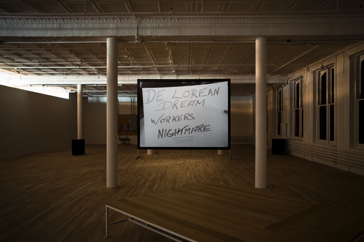 A large room with a wooden table in the foreground. A standing screen is centered between two white poles, showing a projected image of a white sign with handwritten text reading 